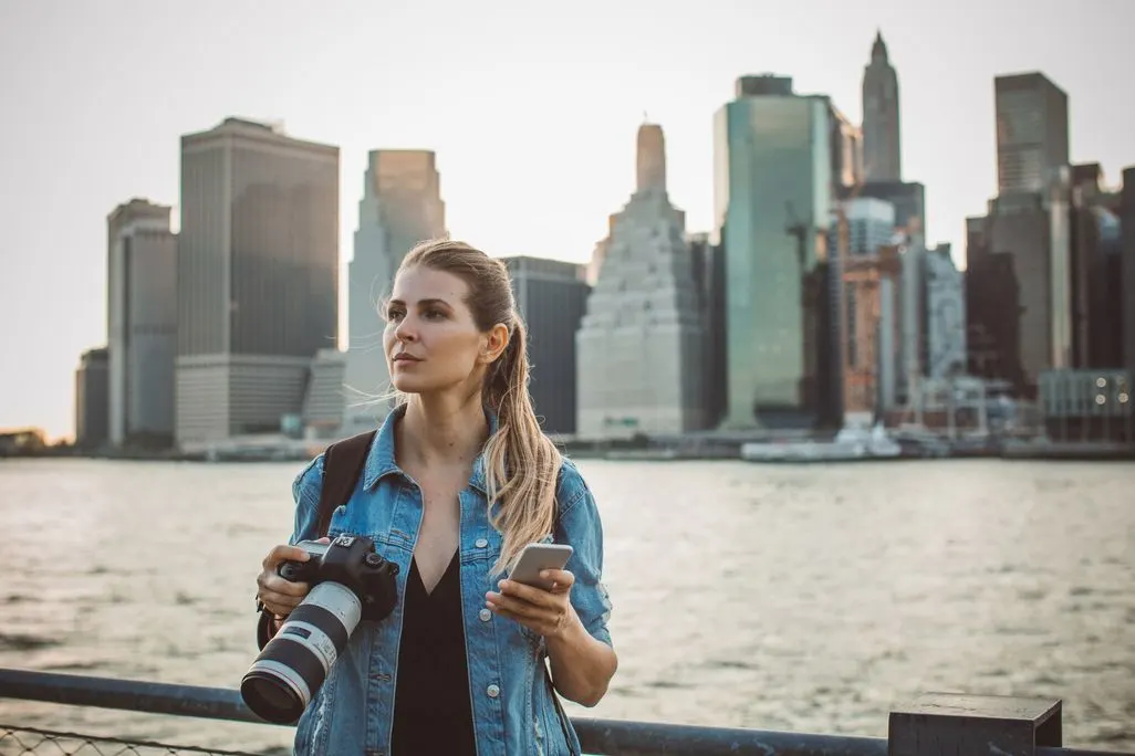 Frau steht mit Kamera und Smartphone vor einer Skyline am Wasser