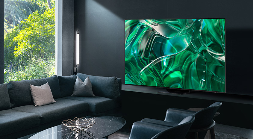 Ein Wohnzimmer mit grauer Couch, Sessel und Tisch. Ein großer Fernseher zeigt abstrakte grüne Formen. Ein Fenster bietet Blick auf Bäume.