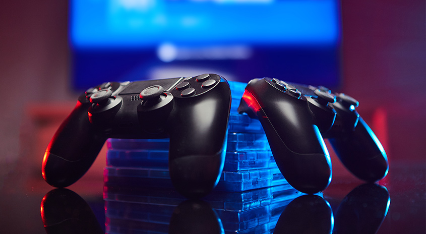 Zwei schwarze Spiel-Controller liegen auf einer Stapel blauer Spielhüllen, vor einem verschwommenen blauen Bildschirm.