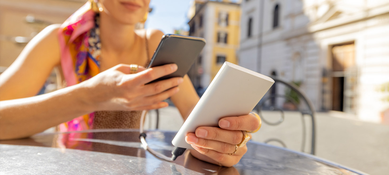 Eine Person hält ein schwarzes Smartphone und eine weiße Powerbank, die mit einem Kabel verbunden sind. Im Hintergrund sind Gebäude zu sehen.