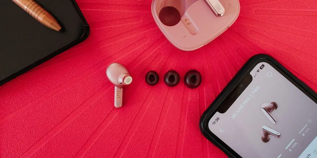 Ein hellbrauner Ohrhörer, drei schwarze Ohrstöpsel, eine offene Ladestation und ein Smartphone liegen neben einem braunen Stift.