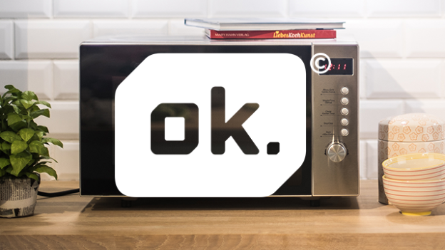 Eine Mikrowelle steht auf einer Holzoberfläche, daneben eine Pflanze und gestapelte Schalen. Bücher liegen oben auf dem Gerät.