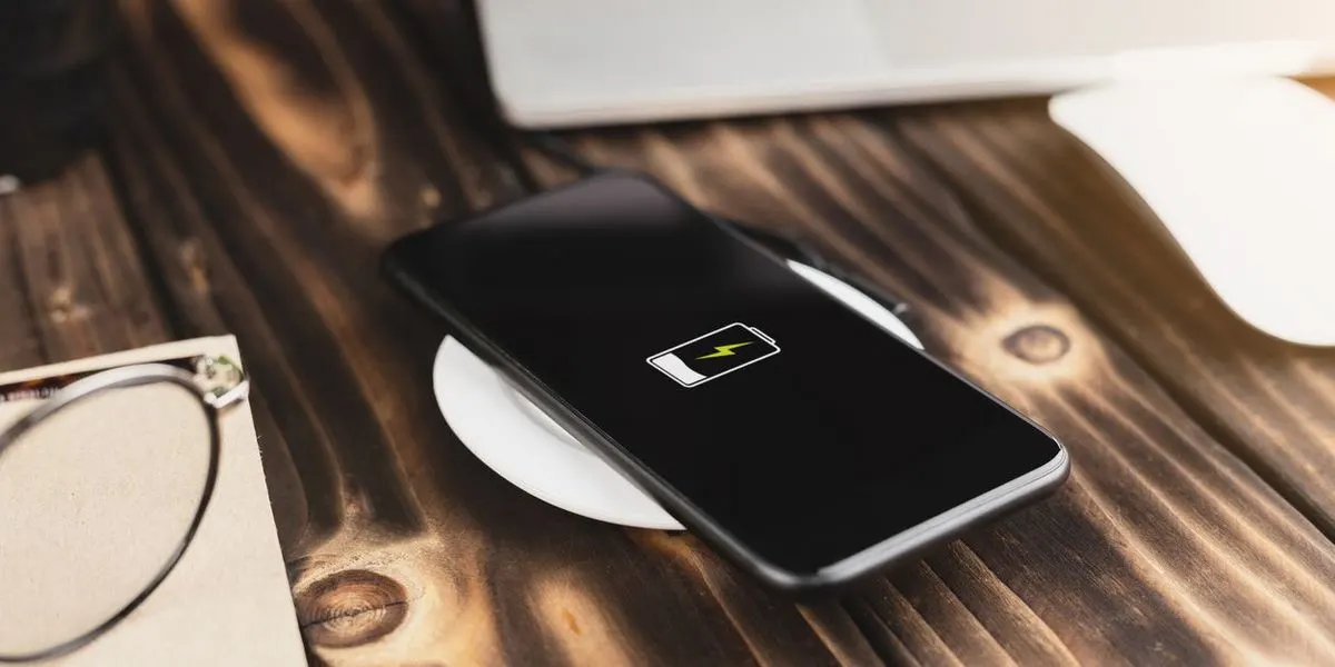 Ein schwarzes Smartphone lädt kabellos auf einem weißen Pad auf einem Holztisch. Im Hintergrund sind ein Laptop, eine Maus und eine Brille zu sehen.