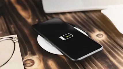 Ein schwarzes Smartphone lädt kabellos auf einem weißen Pad auf einem Holztisch. Im Hintergrund sind ein Laptop, eine Maus und eine Brille zu sehen.