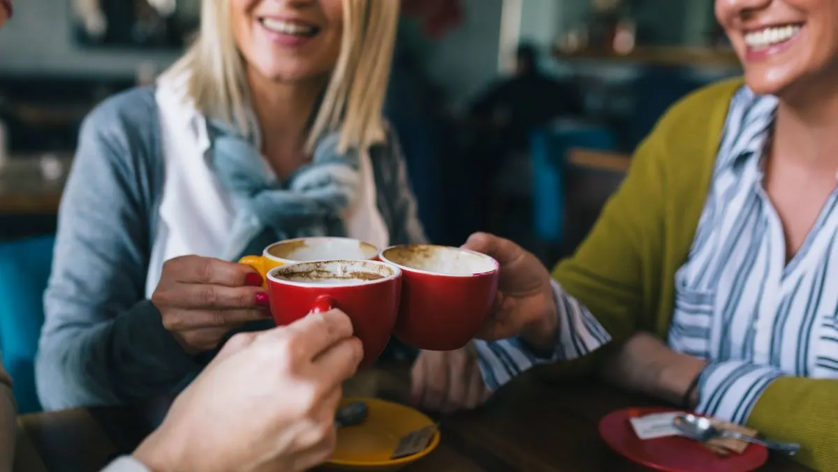 Drei Personen stoßen in einem Café mit zwei roten und einer gelben Kaffeetasse an. Lachende Gesichter, Hände und ein Holztisch sind sichtbar.