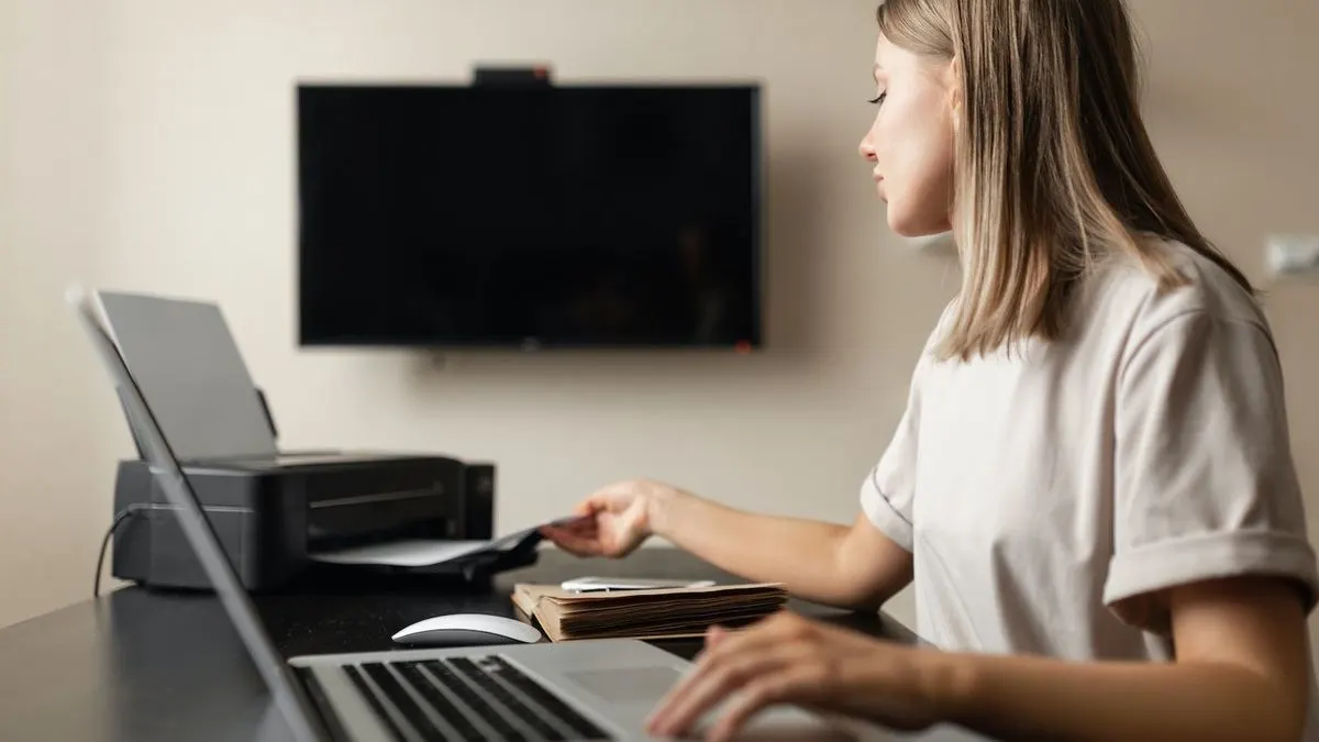 Eine Frau arbeitet konzentriert am Laptop, im Hintergrund ein Fernseher an der Wand.