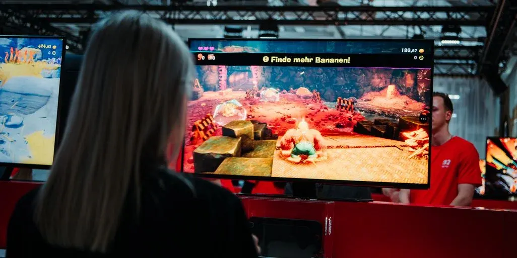 Person spielt an einem Messestand ein Jump’n’Run auf großem Monitor; im Spiel steht „Finde mehr Bananen!“, daneben Betreuer in rotem Shirt.