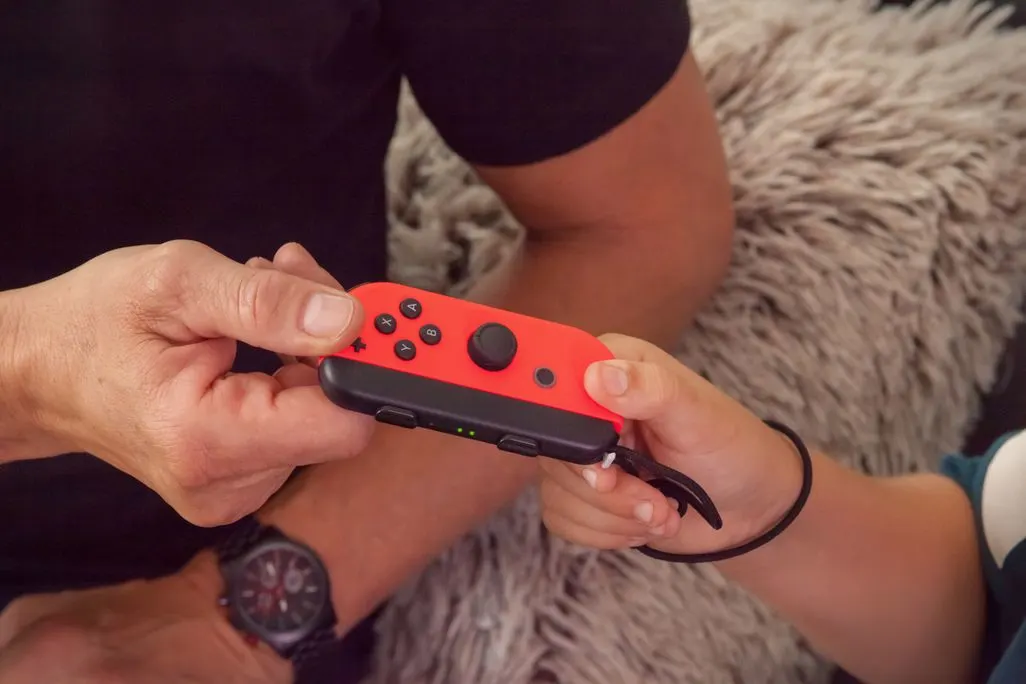 Erwachsene Hand und Kinderhand halten gemeinsam einen roten Nintendo Switch Joy-Con Controller.
