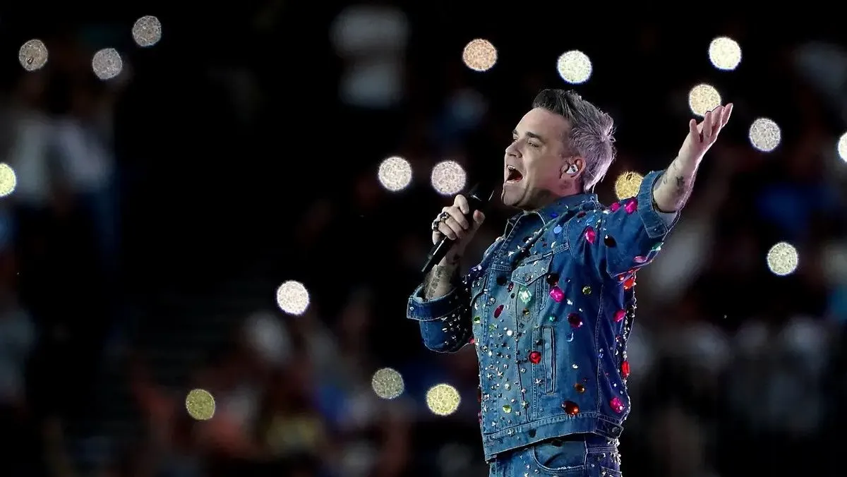 Robbie Williams in blauer Jeansjacke mit bunten Steinen, Mikrofon haltend, singt auf dunkler Bühne mit vielen unscharfen Lichtern.