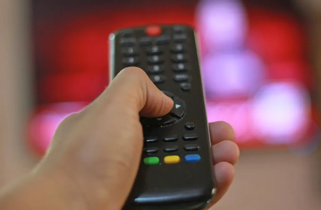 Hand haelt eine Fernbedienung vor einem Fernseher mit rotem Bildschirm.