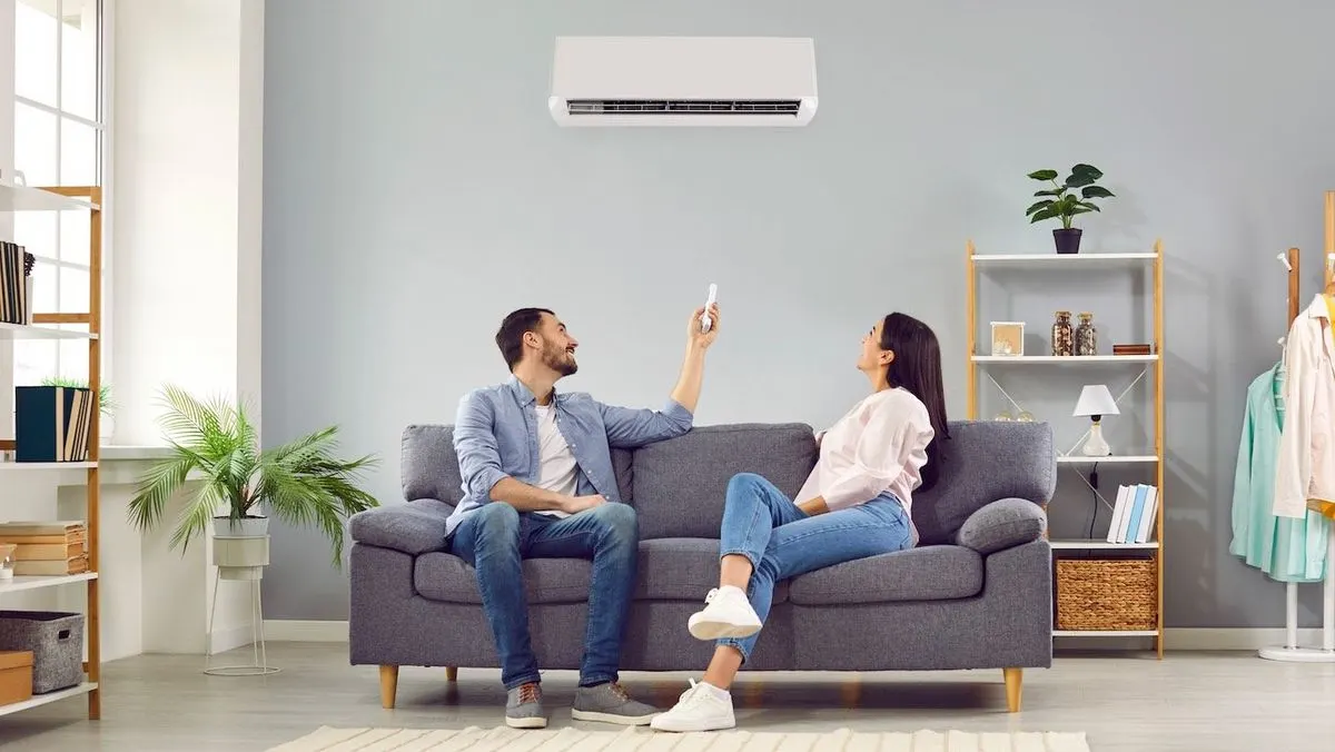 Mann und Frau auf grauem Sofa, Mann zeigt Fernbedienung auf weißes Klimagerät an grauer Wand. Wohnzimmer mit Regalen, Pflanzen, Fenster.