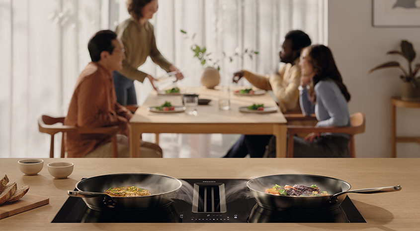 Zwei Pfannen auf einem Kochfeld mit Dunstabzugshaube, ein Tisch mit gedecktem Essen im Hintergrund.