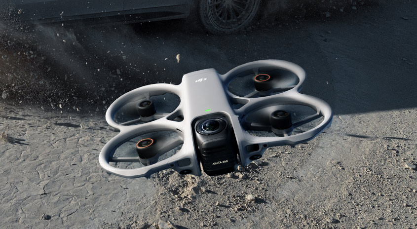  DJI Avata 360 Drohne fliegt knapp über staubigem Boden neben Auto, starke Nahaufnahme der Rotoren und Kamera in rauer Offroad-Umgebung.