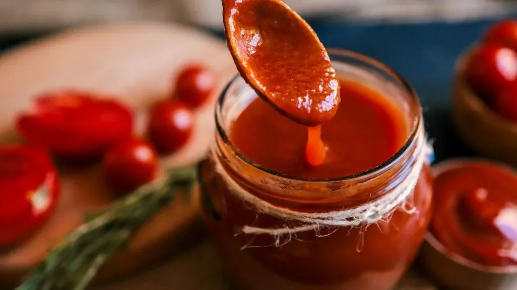 Ein Löffel mit Tomatensauce tropft in ein rustikales Einmachglas, im Hintergrund frische Kirschtomaten und Kräuter auf Holzbrett.