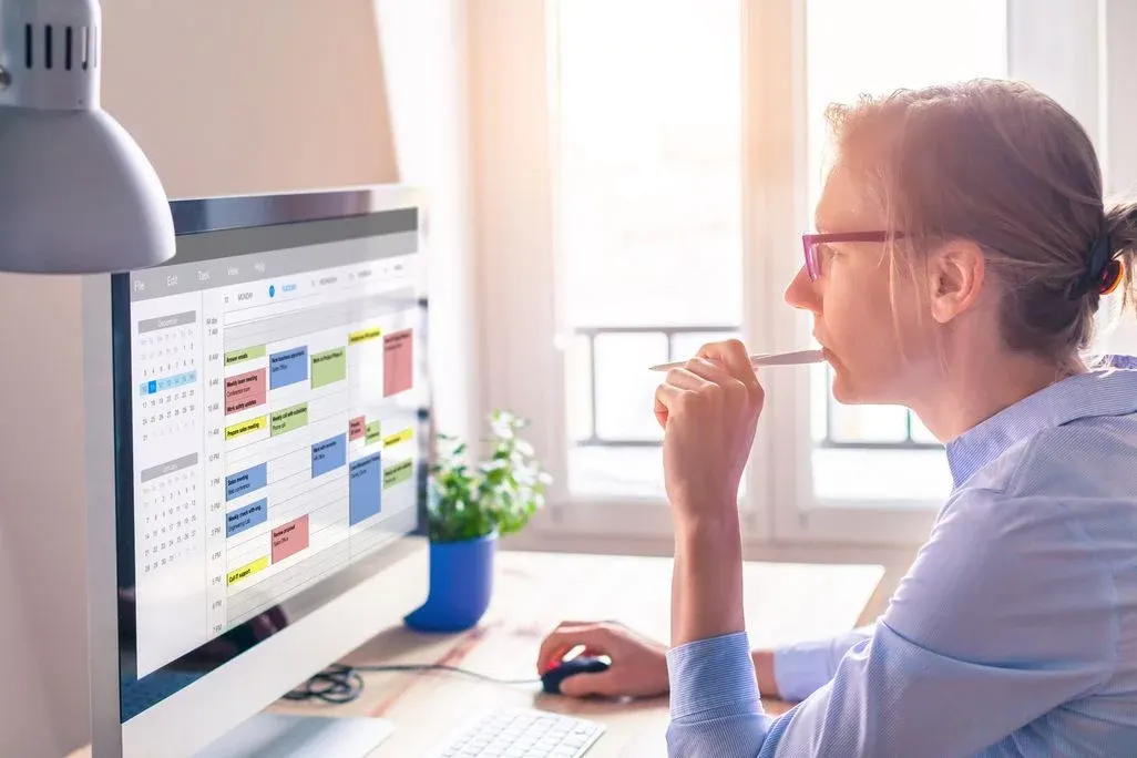 Person mit lila Brille und blauem Hemd blickt auf Monitor mit Terminplaner-Software. Hält Stift am Mund. Schreibtischlampe, Pflanze, sonniges Fenster.