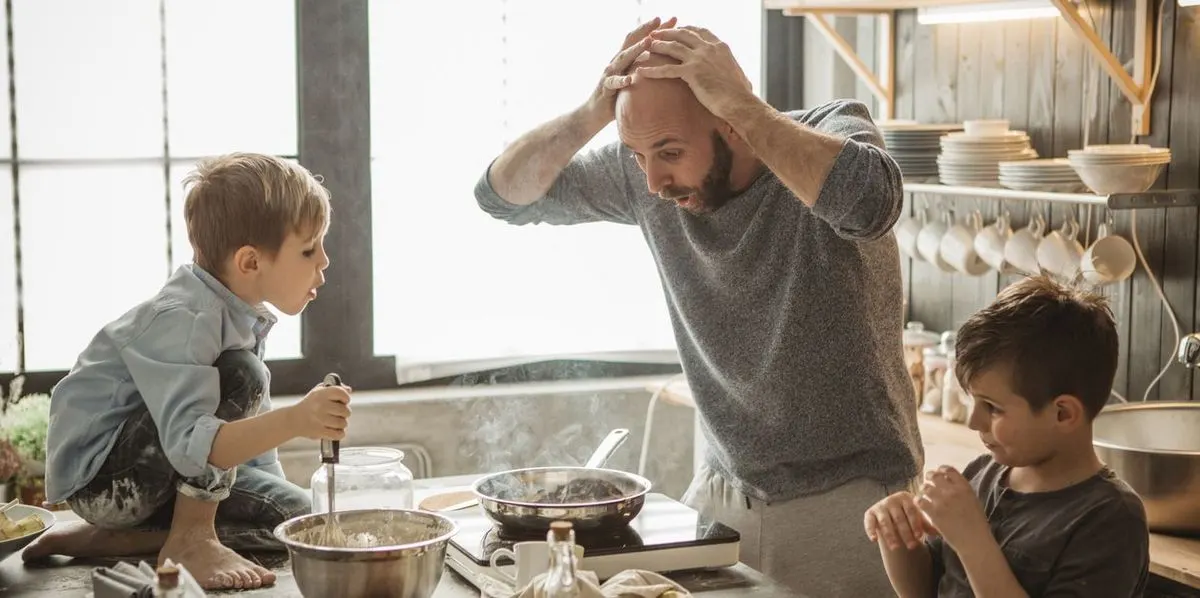 Vater steht in einer Küche und hält sich mit beiden Händen den Kopf, während zwei Kinder helfen zu kochen.