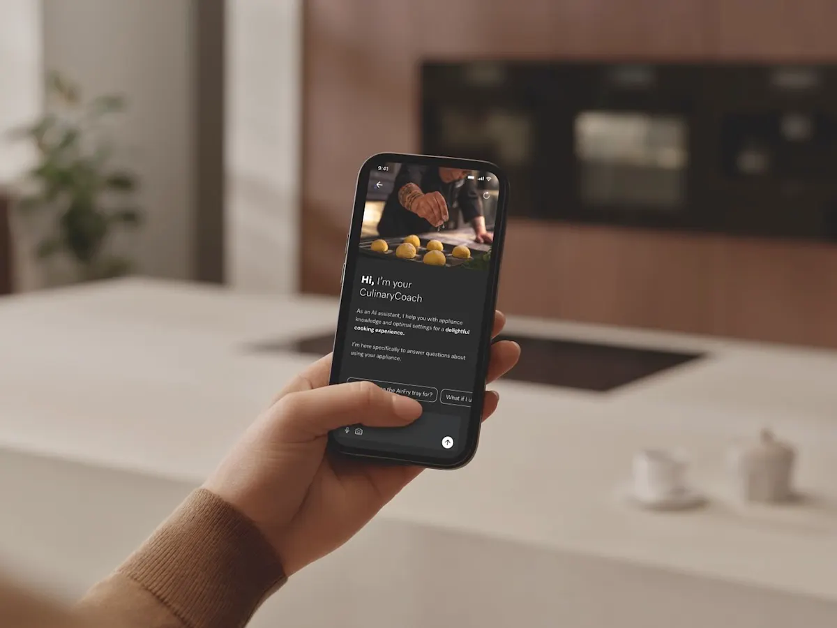 Eine Hand hält ein Smartphone und steuert am Bildschirm den Culinary Coach von Miele. Im Hintergrund ist verschwommen eine Küche mit Backrohr zu sehen.