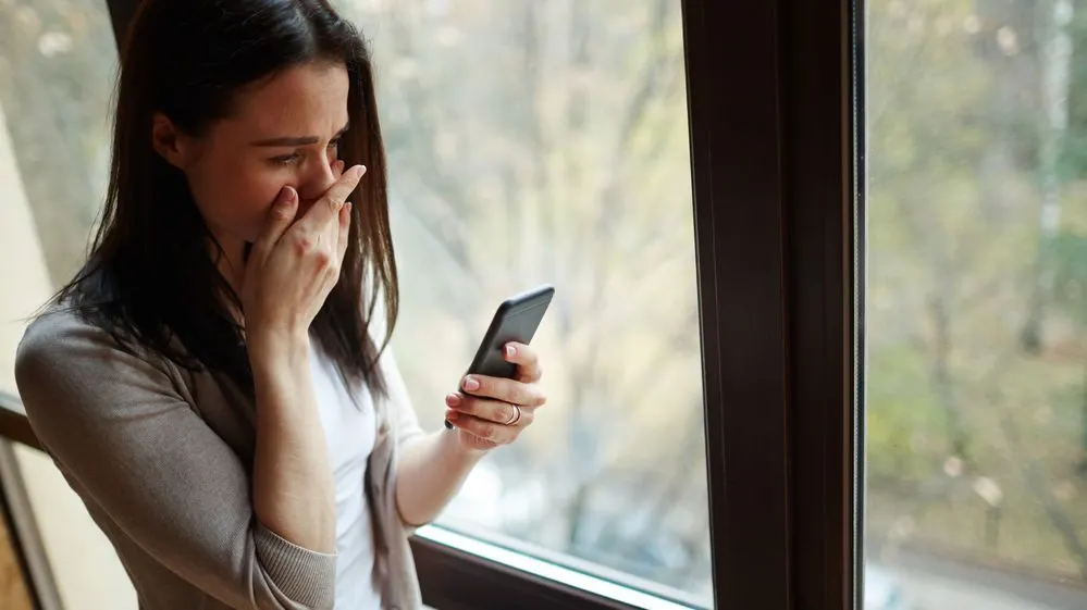 Frau steht am Fenster und schaut verzweifelt auf ihr Smartphone.