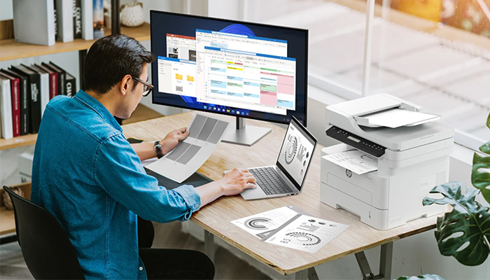 Ein Mann sitzt auf einem Tisch und arbeitet. Auf dem Tisch befindet sich ein HP Monitor, ein Notebook und ein Drucker.