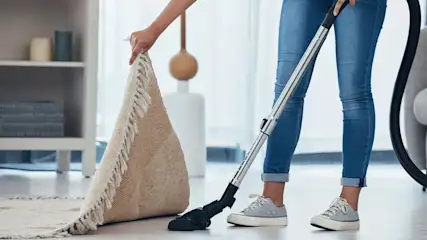 Frau in Jeans hebt einen Fransентeppich an und saugt darunter mit einem Staubsauger den hellen Parkettboden in einem modernen Wohnzimmer.