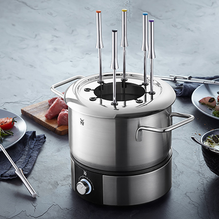 Ein elektrischer Fondue-Topf aus Edelstahl steht auf einem Tisch, umgeben von Tellern, Fleisch und Gabeln.