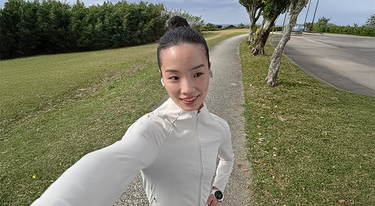 Eine Person macht ein Selfie auf einem Gehweg in einer grünen Parklandschaft. Im Hintergrund sind Bäume, Rasenflächen und eine Straße zu sehen. Die Person trägt eine helle Jacke und eine Smartwatch. Das Gesicht der Person ist unkenntlich gemacht.