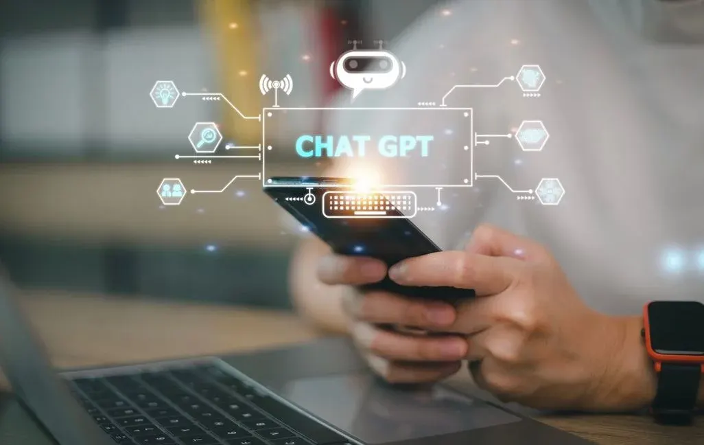 Hände halten ein Smartphone mit leuchtendem 'CHAT GPT'-Overlay, umgeben von KI-Symbolen. Laptop-Tastatur und Smartwatch im Vordergrund.