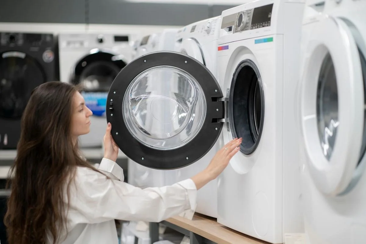Frau mit langen Haaren öffnet die Tür einer weißen Waschmaschine in einem Elektrofachmarkt mit mehreren Geräten im Hintergrund.