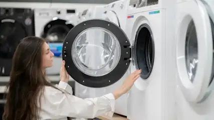 Frau mit langen Haaren öffnet die Tür einer weißen Waschmaschine in einem Elektrofachmarkt mit mehreren Geräten im Hintergrund.