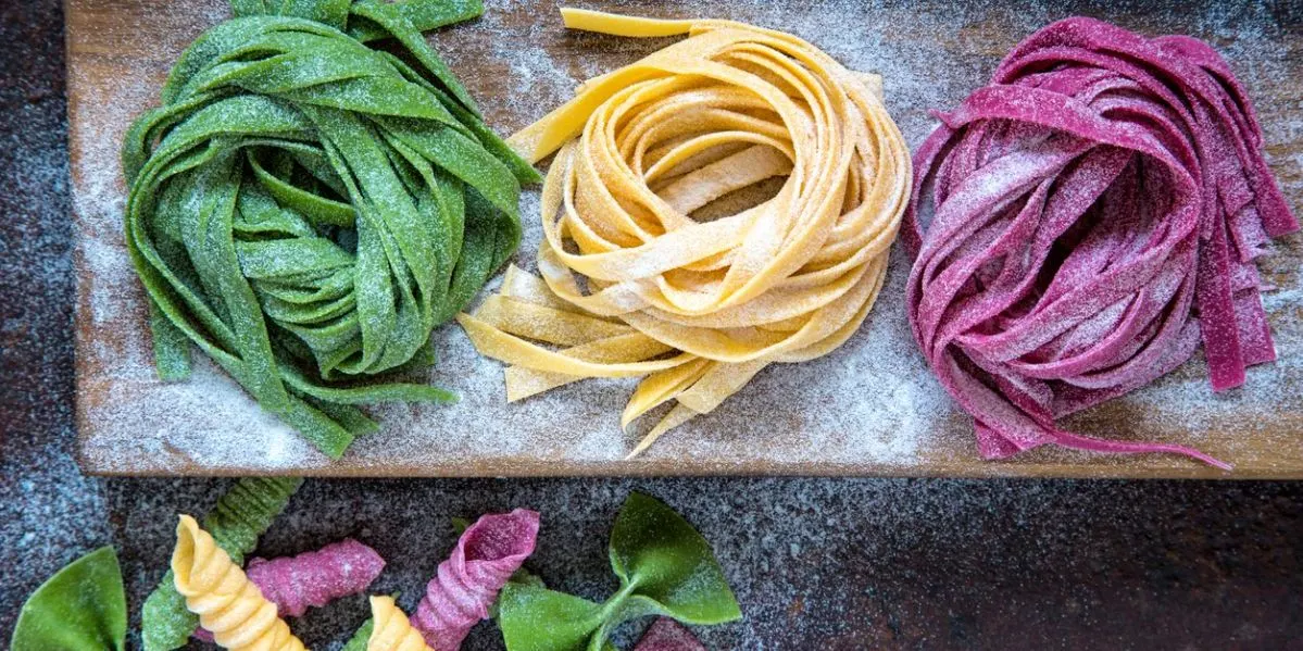 Drei Nester frischer, bemehlter Pasta in Grün, Gelb und Magenta auf einem Holzbrett. Unten liegen lose, bunte Nudeln.