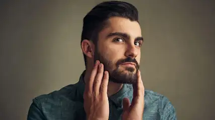 Mann in blauem Denimhemd blickt nach oben rechts. Beide Hände berühren seinen Bart an den Wangen.