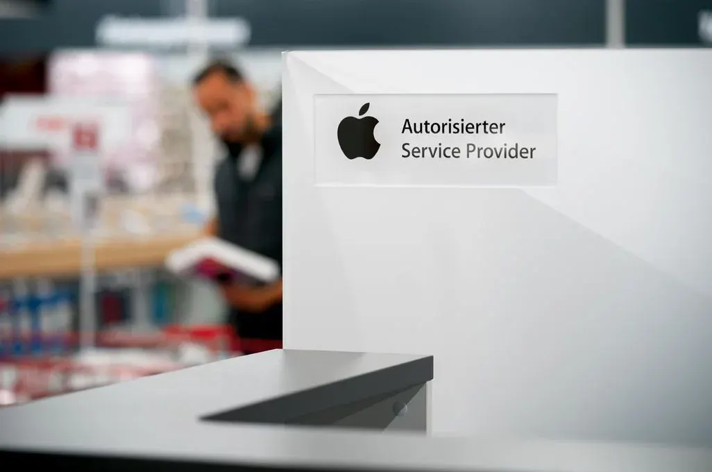 Weißer Tresen mit "Apple Autorisierter Service Provider" Schild. Im Hintergrund hält eine unscharfe Person ein Buch.