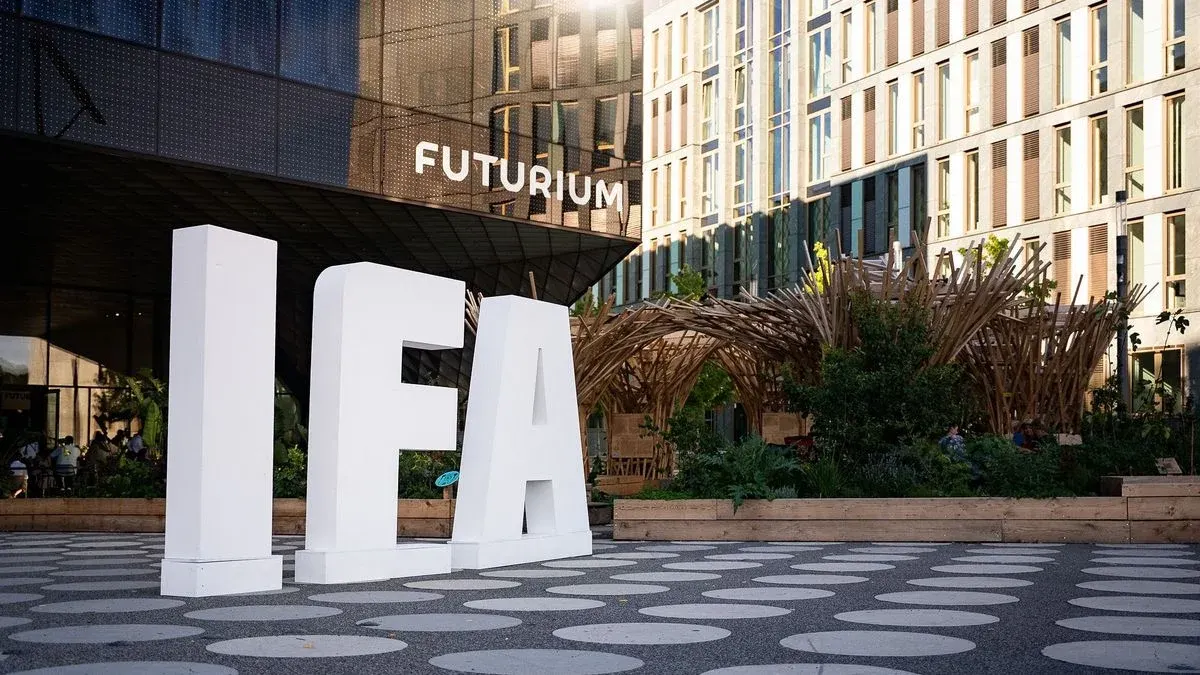 Große weiße IFA-Buchstaben auf gemustertem Boden vor dem dunklen Futurium-Gebäude und hellen Gebäuden mit Pflanzen und Holzstrukturen.