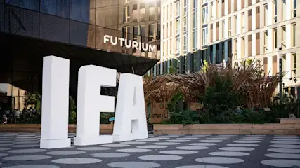 Große weiße IFA-Buchstaben auf gemustertem Boden vor dem dunklen Futurium-Gebäude und hellen Gebäuden mit Pflanzen und Holzstrukturen.