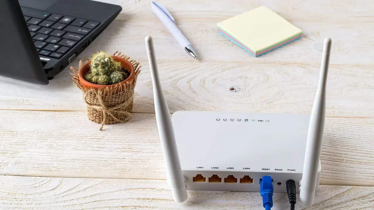 Weißer Router mit zwei Antennen, blauem und schwarzem Kabel. Daneben ein Laptop, Kaktus, Stift und Notizzettel.