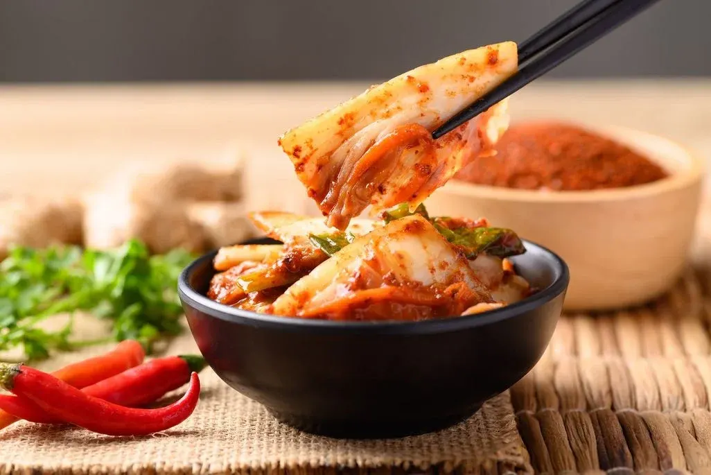Stäbchen heben Kimchi aus einer schwarzen Schale; im Hintergrund rote Chilischoten, frische Kräuter, Ingwer und Gochugaru-Gewürzpulver.
