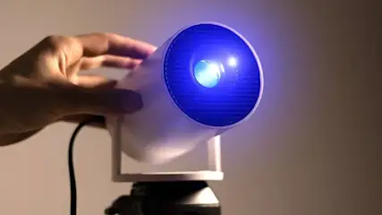 Hand richtet einen modernen, weißen Beamer auf einem Stativ aus, dessen Linse hellblau leuchtet, vor neutralem Hintergrund.