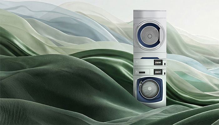 Electrolux Professional Waschmaschine und Trockner im Stack vor wellenförmigem Stoffhintergrund in Grün- und Silbertönen.