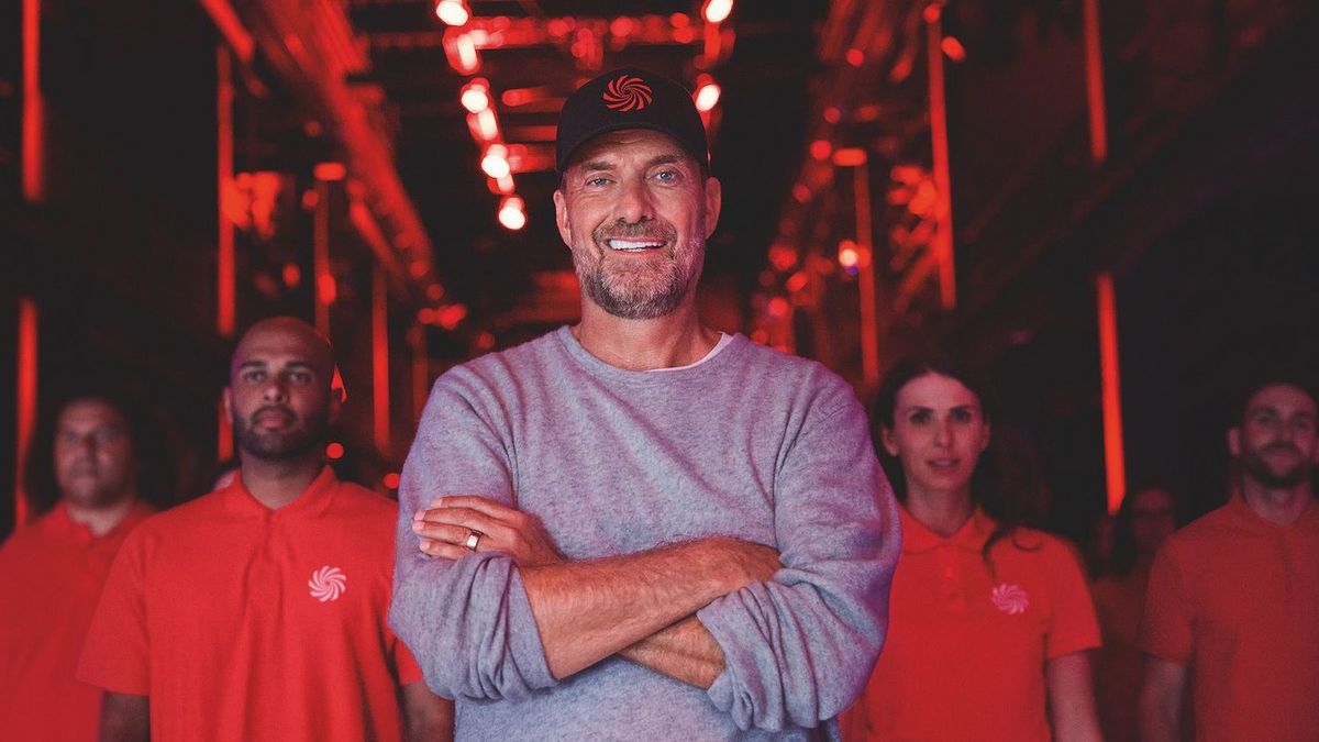 Jürgen Klopp in grauem Pullover und Kappe mit rotem Logo lächelt, Arme verschränkt. Umgeben von Personen in roten MediaMarkt Polohemden in rotem Licht.
