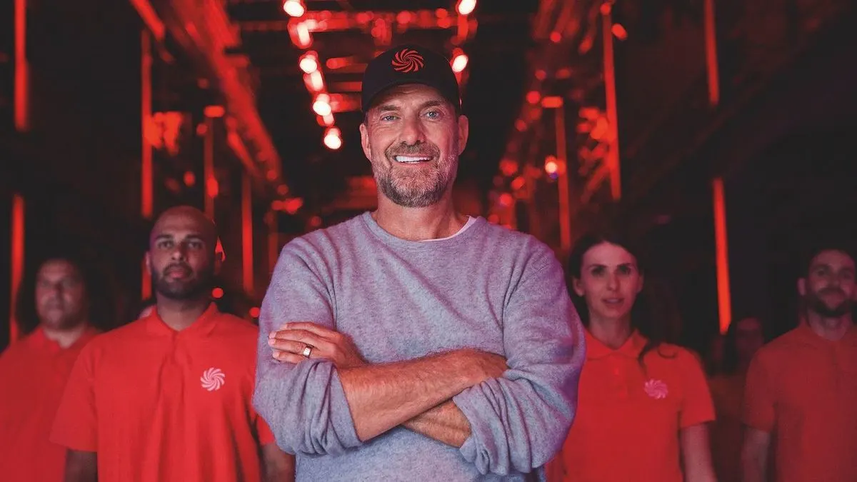 Jürgen Klopp in grauem Pullover und Kappe mit rotem Logo lächelt, Arme verschränkt. Umgeben von Personen in roten MediaMarkt Polohemden in rotem Licht.