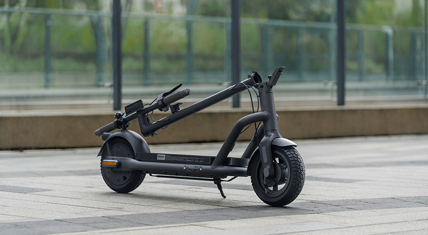 Ein grauer Elektroroller steht auf einem gepflasterten Weg, mit Bäumen und einem Gebäude im Hintergrund.