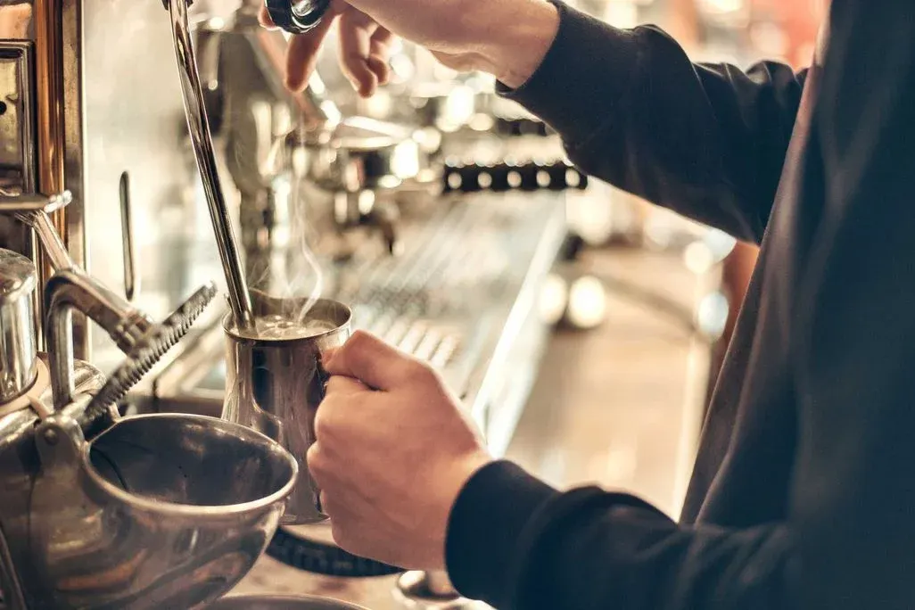 Hände bedienen eine silberne Kaffeemaschine; Dampf steigt aus einem Metallkrug beim Milchschäumen. Glänzende Details im Hintergrund.