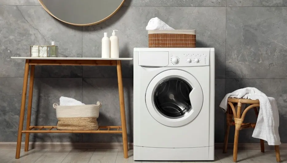 Weiße Waschmaschine, Holztisch mit Spiegel, Rattenschemel, und Handtücher in Körben. Auf hellem Boden vor grauer Fliesenwand.