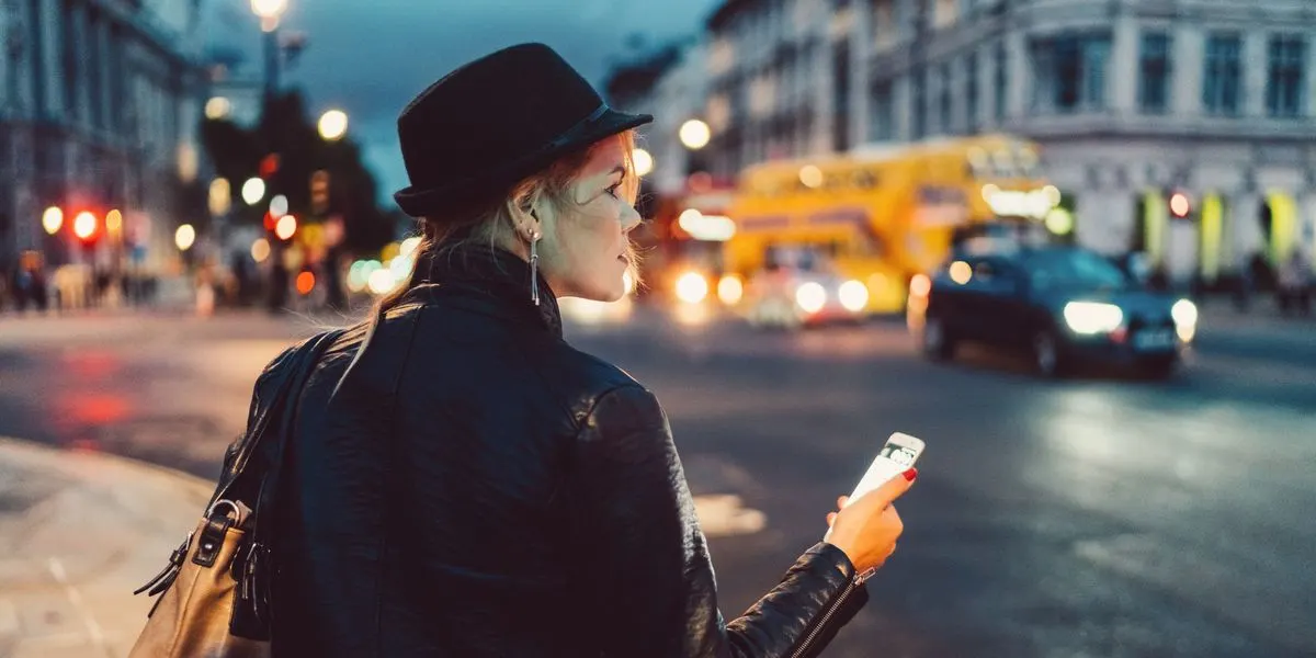 Eine Person mit schwarzem Hut, Lederjacke und brauner Tasche hält ein weißes Smartphone. Rote Nägel und silberne Ohrringe.