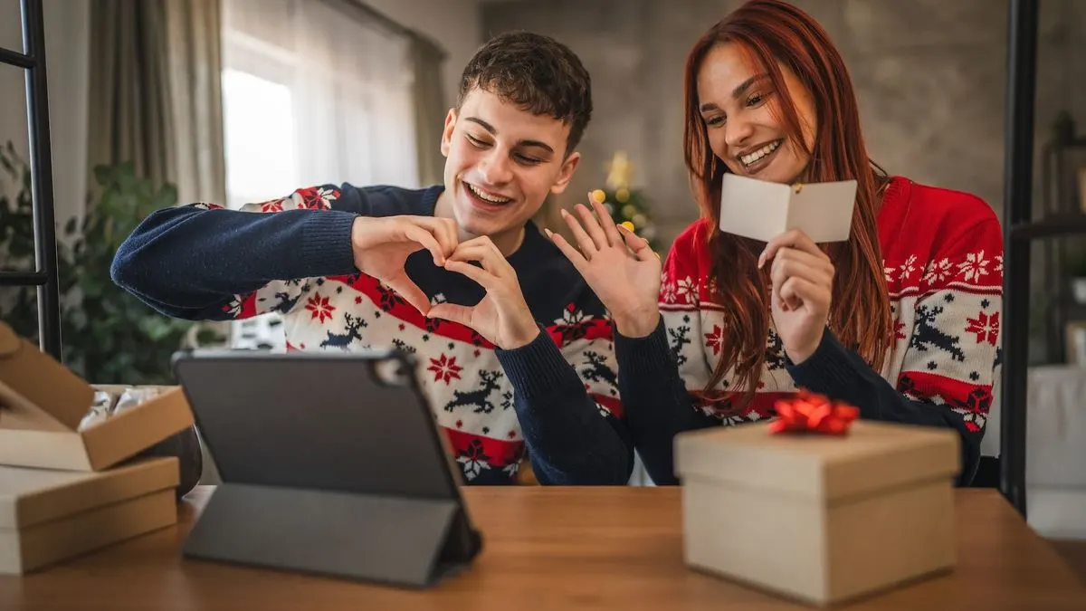 Zwei Personen in festlichen Pullovern sitzen am Tisch mit Geschenkboxen und halten gemeinsam ein Herz mit den Händen vor einem Tablet.