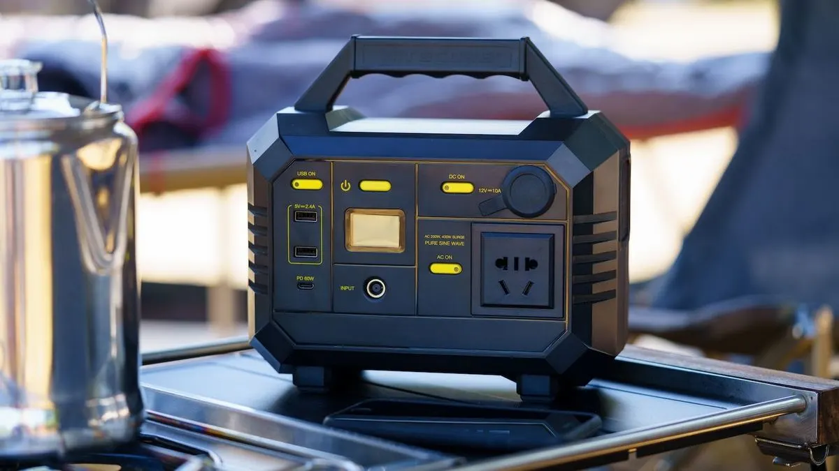 Schwarze tragbare Powerstation mit gelben Anschlüssen und Display auf einem Metalltisch. Links ein silberner Kessel, im Hintergrund Camping.