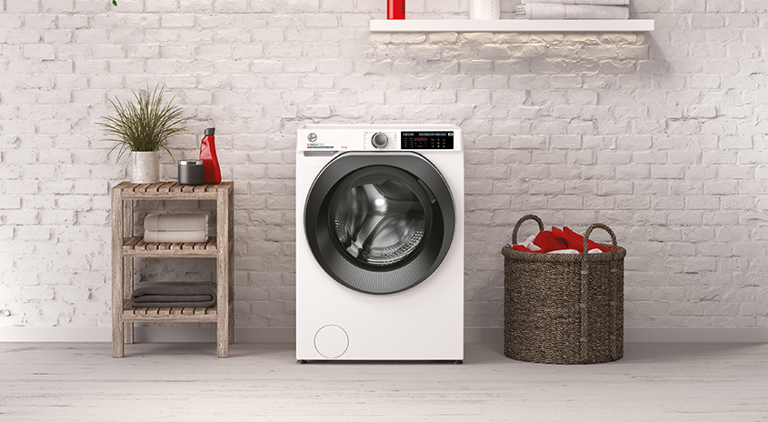 Im Vordergrund steht eine weisse Waschmaschine mit dunkler Trommel, daneben ein Wäschekorb und ein Regal. Backsteinwand im Hintergrund.
