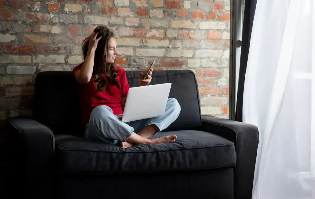  Person sitzt entspannt barfuß auf einem dunklen Sofa vor Backsteinwand, Laptop auf dem Schoß, Smartphone in der Hand, Licht fällt durch ein großes Fenster.