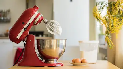 Rote Kitchen Aid Küchenmaschine mit Schneebesen und weißer Masse über Metallschüssel. Daneben Eier, Mehlbehälter auf Holztisch. Gelbe Mimosenblüten im Hintergrund.