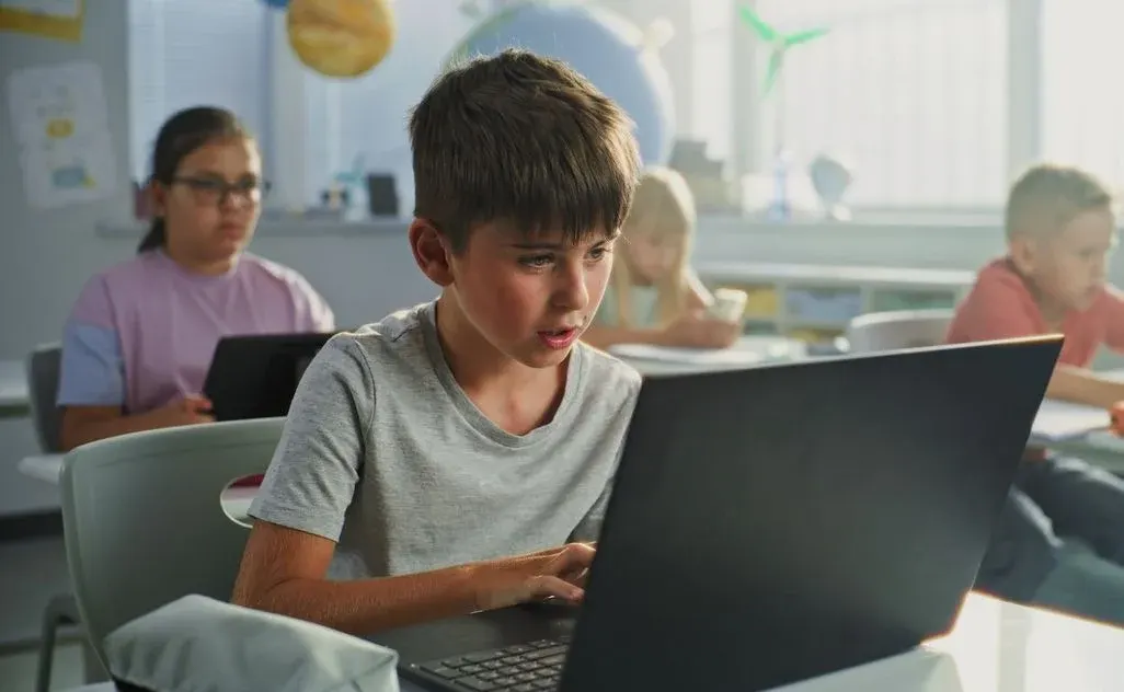 Volksschüler konzentriert am Laptop im Klassenraum, Mitschüler im Hintergrund und Lernmaterialien sichtbar.